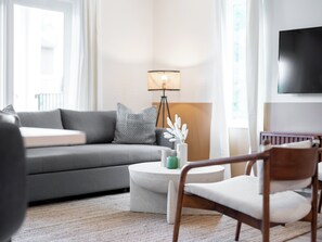 Ein modernes Wohnzimmer mit einem grauen Sofa, einem runden Couchtisch, einem Holzstuhl, einer Stehlampe und einem an der Wand befestigten Fernseher.