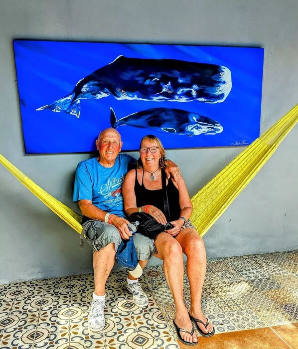 Deux personnes sont assises sur un sol à motifs, près d’un hamac, d’un tableau bleu représentant une baleine et d’un mur recouvert de carreaux.