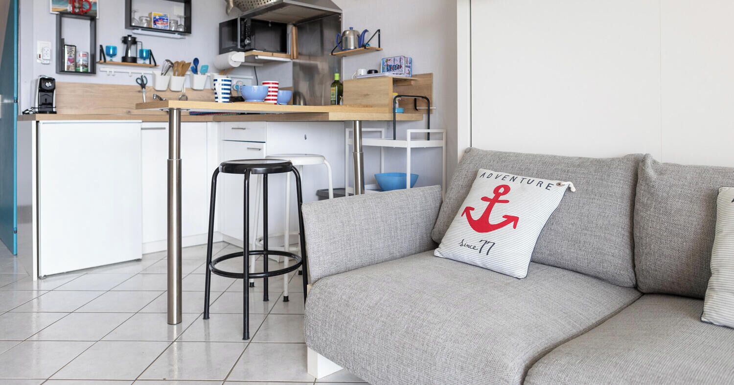 A modern kitchen with a dining area, a grey sofa with a cushion, and a bar stool.