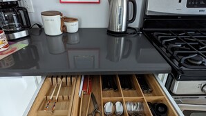 Eine Küchenarbeitsfläche mit einer Kaffeemaschine, Zucker- und Kaffeedosen sowie einem Wasserkocher. Darunter befindet sich eine Schublade mit Besteck wie Löffeln, Gabeln und einer Schere.