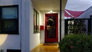 Ein Gebäudeeingang mit roter Tür, einer kleinen Veranda und einem Gartenbereich mit Pflanzen.