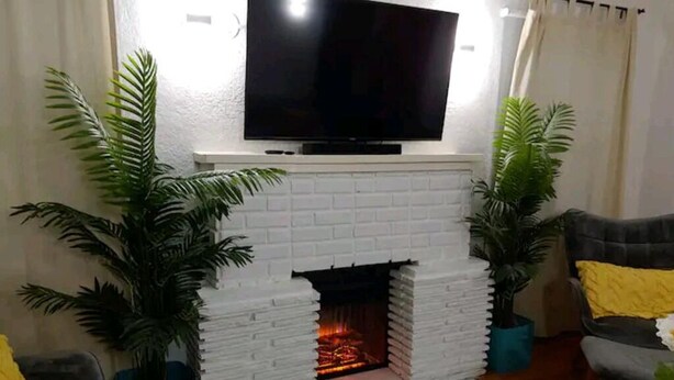 Ein Wohnzimmer mit Kamin aus weißem Mauerwerk, einem flachbildfernseher darüber und je einer Topfpflanze auf jeder Seite.