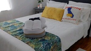 Ein ordentlich bezogenes Bett mit einem Bettbezug mit tropischem Blattmuster, ein weißer Wäschekorb und ein Kissen mit dem Kalifornien-Wappen.