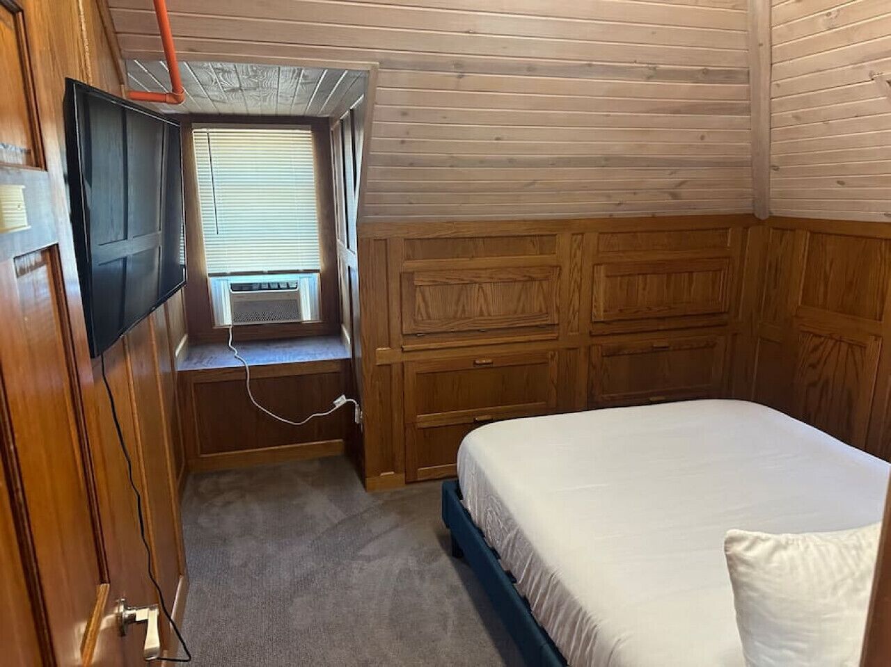 Ein kleines, gemütliches Schlafzimmer mit einem Einzelbett, einem an der Wand montierten Fernseher und einem Fenster mit Jalousien.