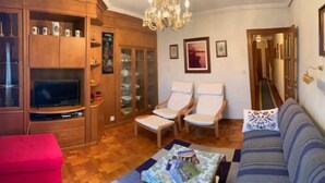 Un salon comprenant un téléviseur, deux fauteuils, un canapé, un pouf rouge et des meubles en bois.