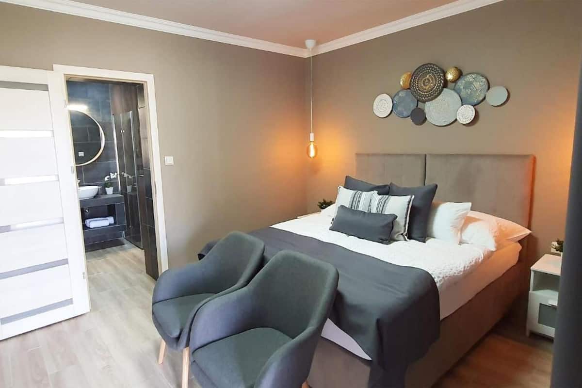 A bedroom with a bed, two chairs, a bedside table, a wall decoration, and a door leading to a bathroom.
