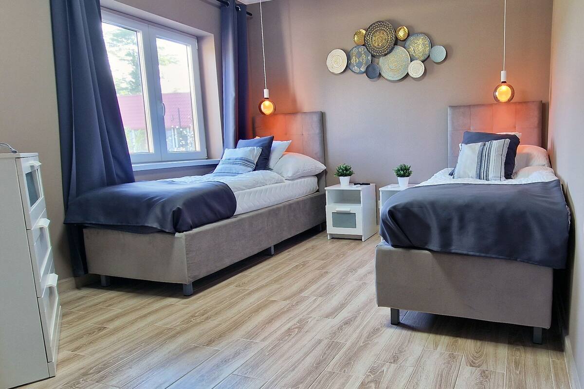 A bedroom with two beds, a window with curtains, a bedside table with a plant, and a wall decoration.