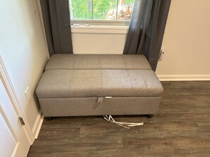 A grey upholstered bench in a room with wooden flooring, a window with curtains, and a white wall. - The Bear Cave - Jasper GA (Jasper)