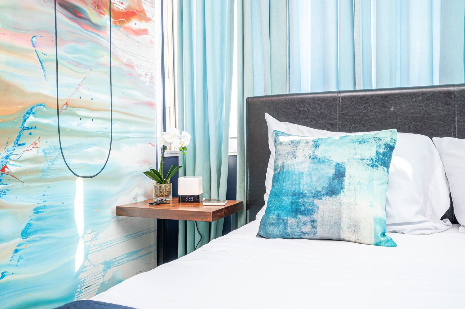 A hotel room with a bed, a nightstand with a clock, a vase with flowers, and a wall with abstract art.