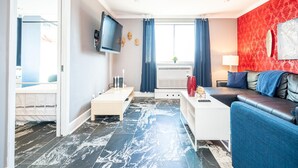 A modern living room with a sofa, a coffee table, and a TV mounted on the wall. There is a window with curtains, a small table with a vase, and a red wall with a decorative item. - ATL Downtown Condo with Free Parking (Atlanta)