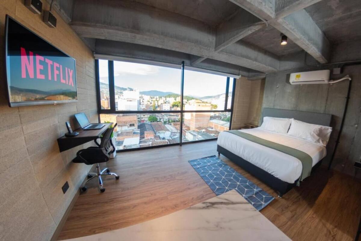 A modern hotel room with a large bed, a desk with a laptop, a TV mounted on the wall, and a large window offering a city view.