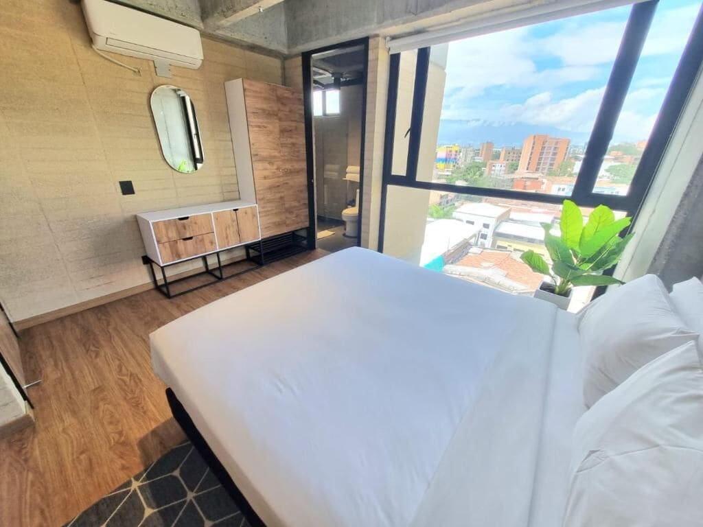 Un dormitorio moderno con una cama grande, un tocador de madera, un espejo y vistas a la ciudad a través de un ventanal.