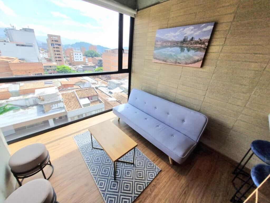 Quarto com sofá, tapete estampado, banqueta de bar e vista da cidade através de grandes janelas.