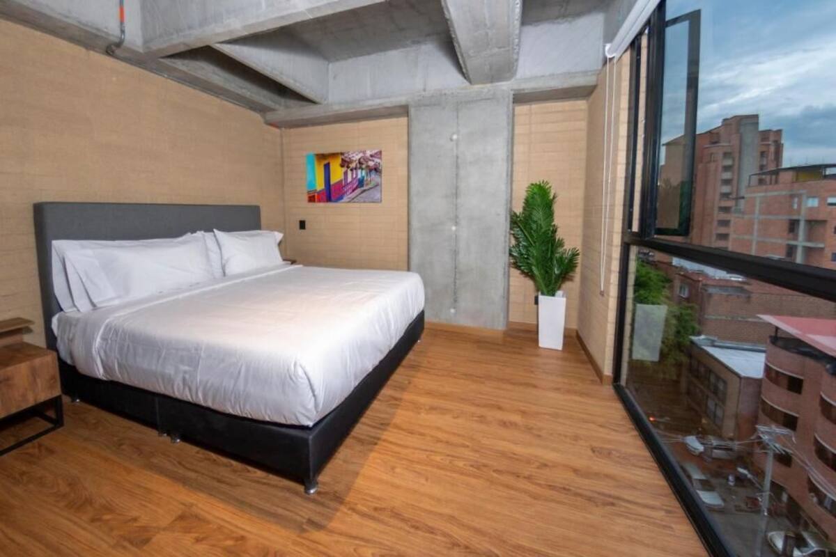 A modern bedroom with a large bed, a potted plant, and a city view through a window.