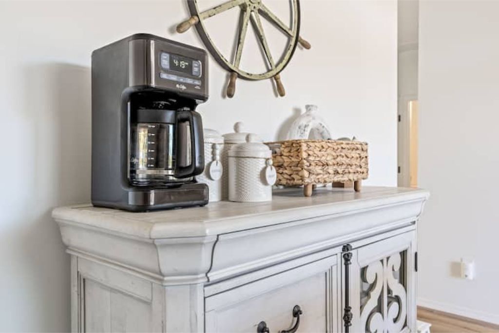 A coffee maker on a white cabinet with a decorative wheel and storage containers.