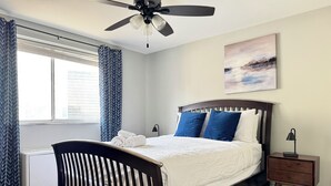 Ein Schlafzimmer mit Bett, Nachttisch, Deckenventilator und einem Fenster mit Vorhängen.