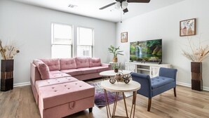 A living room with a pink sofa, a blue chair, a small round table, a ceiling fan, and a television on a white cabinet. - La Vida Loca Stay in Baton Rouge near LSU - BC1011 (Baton Rouge)