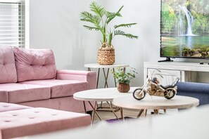 Moderni olohuone, jossa on vaaleanpunainen sohva, pyöreä sohvapöytä, pieni sivupöytä, jossa on kasvi, ja televisio, josta näkyy luontokuva.