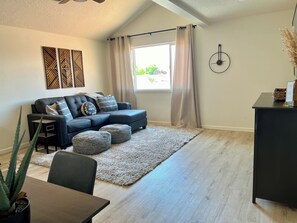 Ein modernes Wohnzimmer mit Ecksofa, Esstisch und großem Fenster mit Vorhängen.