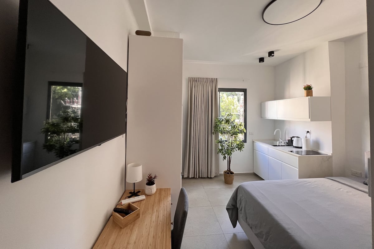 A modern hotel room with a large bed, a flat-screen TV mounted on the wall, a bedside table with a lamp, a small plant, and a view of the outdoors through a window with curtains.