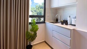 A modern kitchen with white cabinets, a countertop, and a window with beige curtains. - White Door Apartments (Haifa)