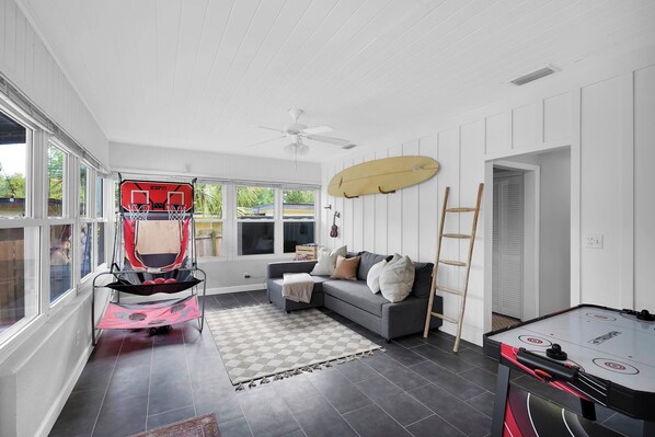 A spacious room with a grey sofa, a red and black baby stroller, a table hockey game, and a surfboard on the wall.