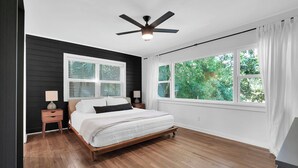 A bedroom with a bed, a nightstand, a lamp, a ceiling fan, and a large window with curtains.