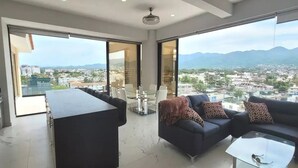 Ein großzügiger Wohnbereich mit einem großen Glasschiebefenster zum Balkon, einer dunklen Sofagarnitur, einem Glastisch und einer modernen Küchenzeile.