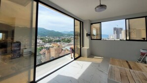 Ein geräumiger Balkon mit großen, raumhohen Fenstern und Blick auf die Stadt, ein Holztisch und ein Deckenventilator.