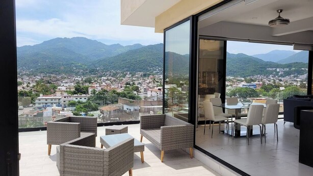 Ein moderner Außenbereich mit Korbmöbeln und einer Glasabtrennung, mit Blick auf eine Stadtlandschaft und dahinter Berglandschaft.