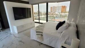 Ein modernes Schlafzimmer mit einem großen Bett, einem an der Wand montierten Fernseher und Blick auf die Stadt durch große Fenster.