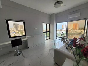 Een moderne woonkamer met een marmeren vloer, een zwarte stoel, een glazen salontafel en een bank. Grote ramen bieden uitzicht op een stadsgezicht.
