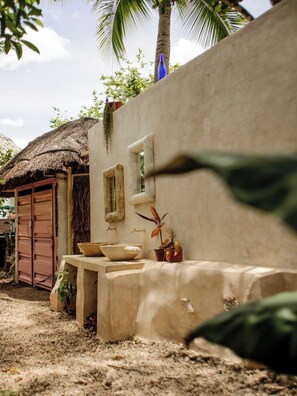 Un entorno rústico al aire libre con una construcción de techo de paja, un muro de hormigón y una pequeña mesa con un cuenco y plantas en macetas.
