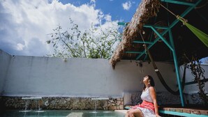 A woman sitting by a pool with a thatched roof structure and a stone wall in the background. - Aldea Toktli Hotel and Museum - Pool, Starlink Wi-Fi, and Mayan House Museum Tour (Izamal)