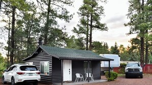 Une petite cabane avec une véranda, deux chaises et une voiture blanche garée devant.