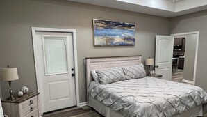 A bedroom with a bed, a ceiling fan, a painting, and a door leading to a kitchen.
