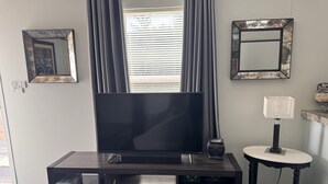A living room with a flat-screen TV on a dark wooden TV stand, a small side table with a lamp, and a window with blinds. - Charming Mountain Retreat Near Grayson Highlands, 2BR 2BA, Sleeps 6 (Mouth of Wilson)