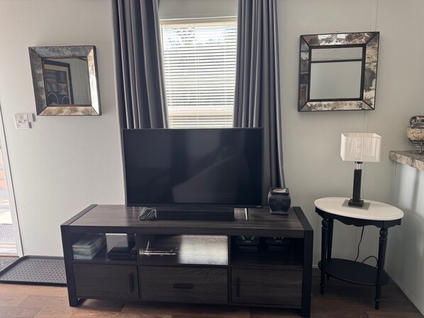 A living room with a flat-screen TV on a dark wooden TV stand, a small side table with a lamp, and a window with blinds.