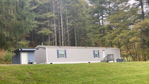 A single-story mobile home with a small attached garage, situated in a grassy area with dense trees in the background. - Charming Mountain Retreat Near Grayson Highlands, 2BR 2BA, Sleeps 6 (Mouth of Wilson)