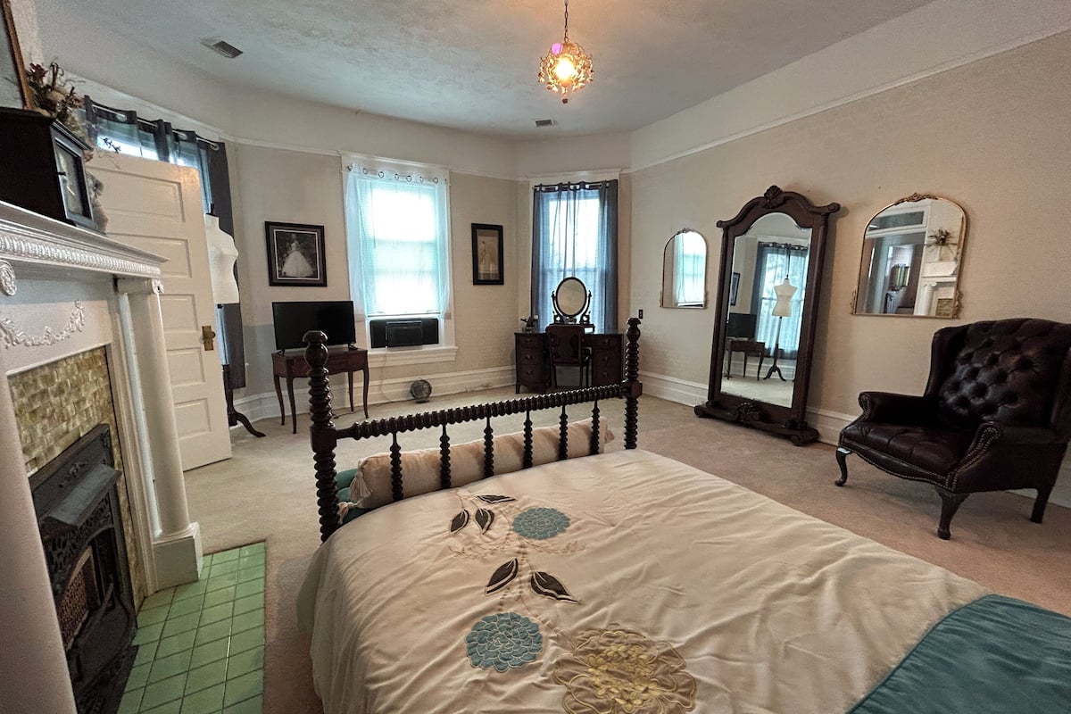 A bedroom with a large bed, a dresser, a mirror, and a fireplace.