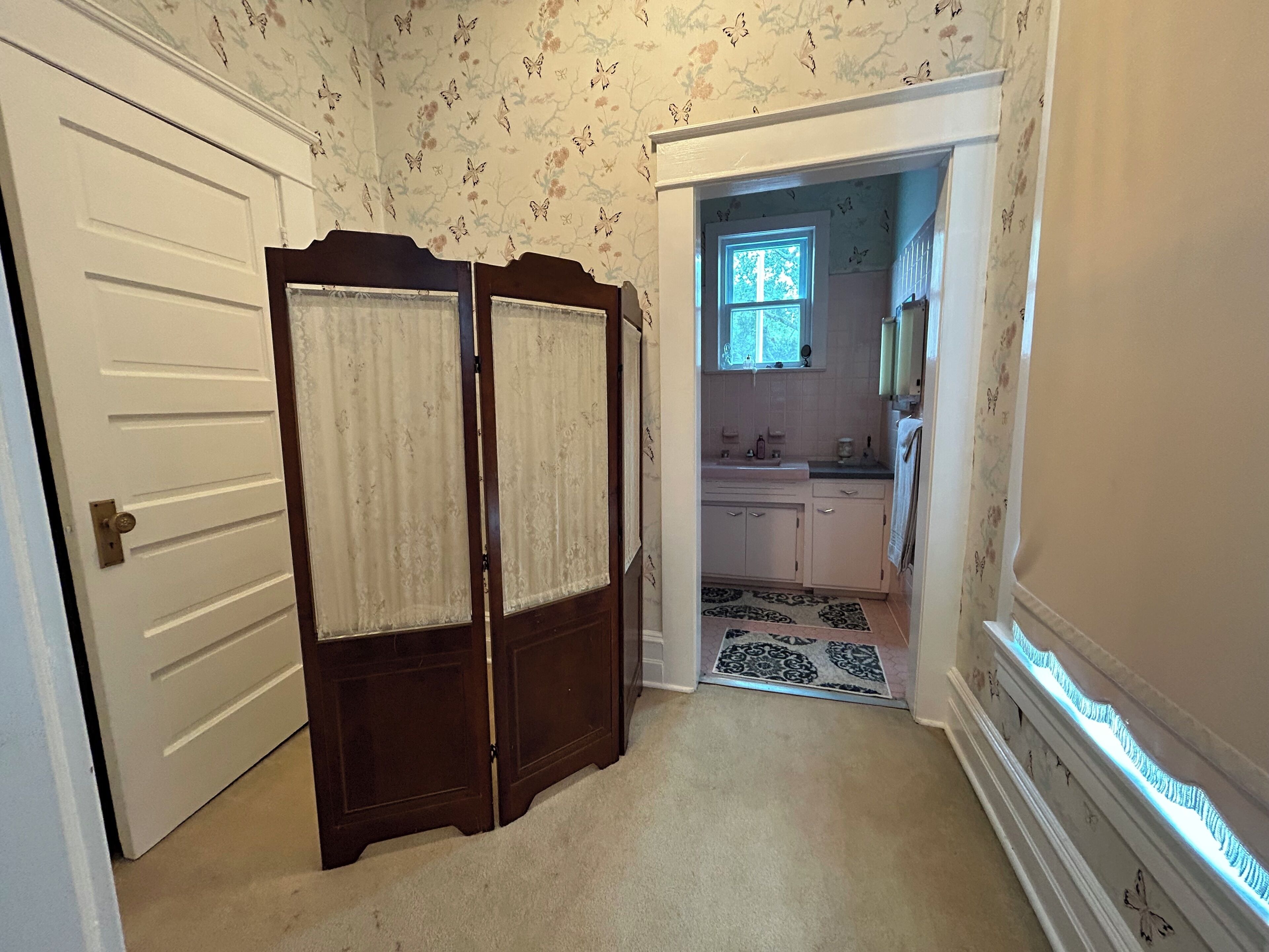 A hallway with a wooden door, a patterned carpet, and a view into a kitchen with white cabinets and a window.