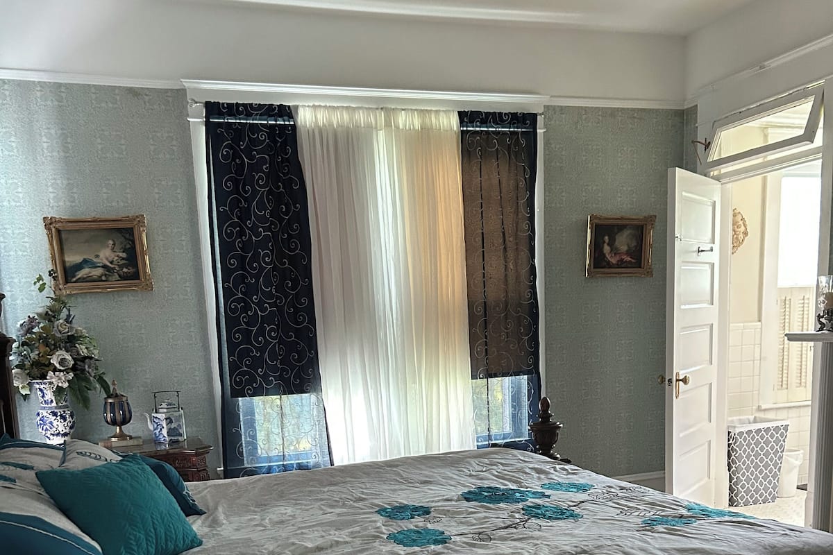 A bedroom with a large bed, a nightstand, a dresser, a window with curtains, and a door.