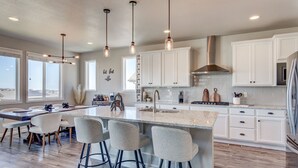 Cocina moderna con armarios blancos, una isla grande con taburetes altos, iluminación colgante y una campana extractora de acero inoxidable.