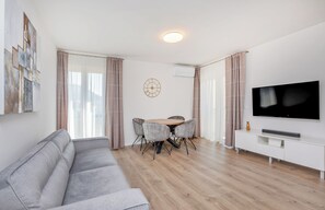 Un salon moderne avec un canapé gris, une table à manger en bois et quatre chaises, un téléviseur à écran plat posé sur un meuble blanc, et un grand tableau abstrait accroché au mur.