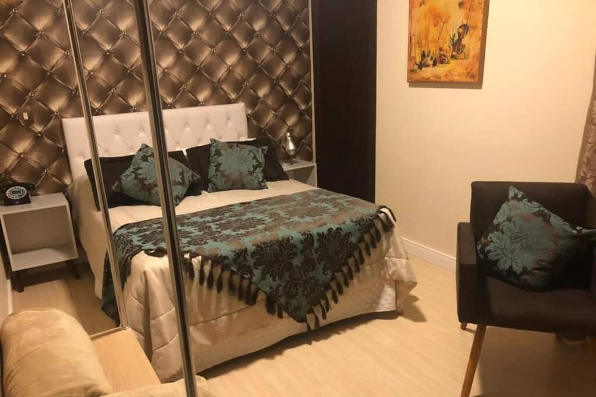A bedroom with a tufted headboard, a bed with patterned bedding, a bedside table with a lamp, a chair with a cushion, and a painting on the wall.