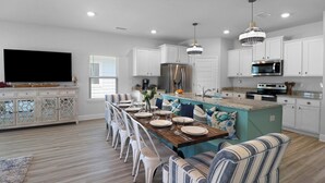 A modern kitchen with white cabinetry, a large island with blue accents, and a dining area with striped chairs. - Worth the Coast Beach House-Golf Cart, Pool, In-law suite, resort community+more (Port St. Joe)