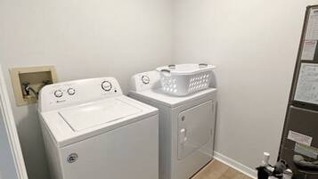 A laundry room with a front-loading washer and dryer, a drying rack, and a small shelf with cleaning supplies.