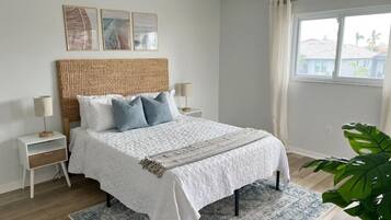 A bedroom with a large bed, a bedside table, a nightstand, a window with curtains, and a plant.