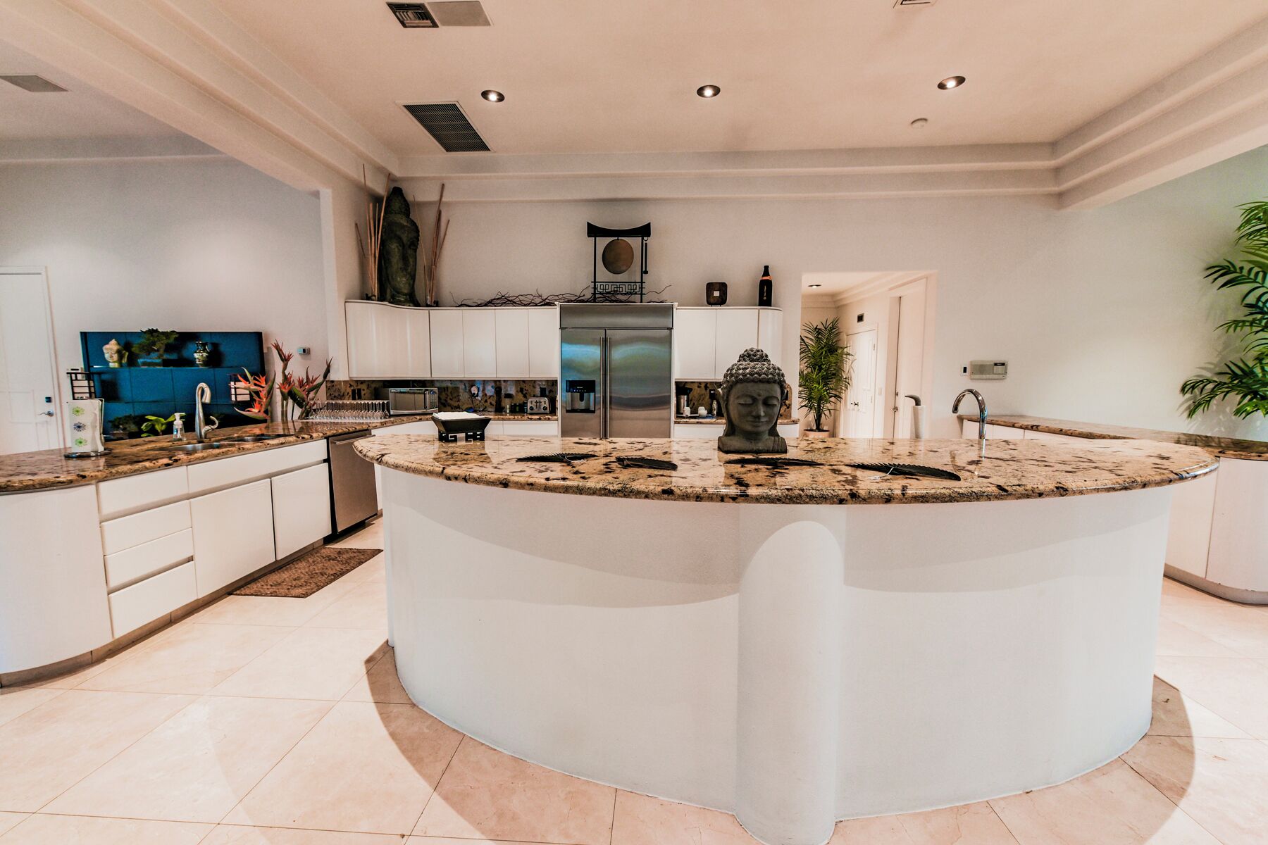 Uma cozinha moderna com uma ilha central, armários brancos, eletrodomésticos de aço inoxidável e uma estátua decorativa de Buda.