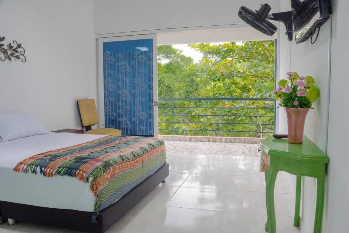 A bedroom with a bed, a green side table, a blue door, and a balcony with a view of greenery.